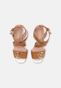 Ruskeat nahkasandaalit kiiltävällä hopeanvärisellä pohjalla, joissa on kaksi sivuttain vinottain kulkevaa hihnaa ja kultainen kuusikulmainen koriste varvashihnassa.