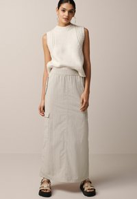 Haut sans manches en tricot blanc avec une texture côtelée, associé à une jupe maxi grise claire avec des poches latérales et une taille élastique. Sandales noires à brides.