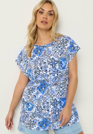 Kobieta w luźnej, niebiesko-białej bluzce w kwiaty z krótkim rękawem oraz jasnych jeansowych szortach, stojąca na jednolitym białym tle.