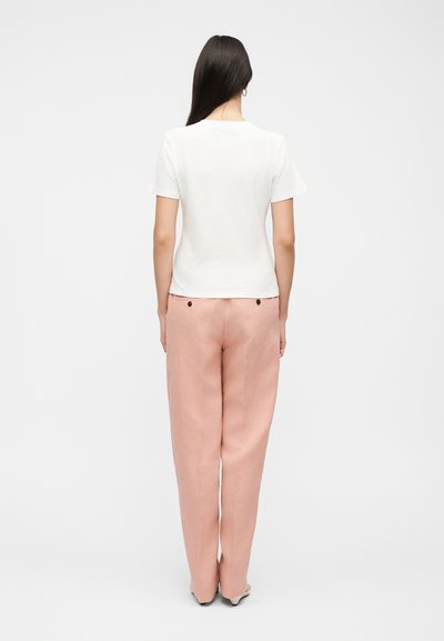 Γυναίκα με μακριά σκούρα μαλλιά στέκεται με την πλάτη, φοράει λευκό κοντομάνικο τοπ και φαρδιά ροζ παντελόνια με κουμπιά στο πίσω μέρος.