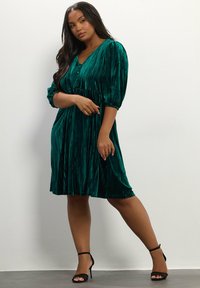 Grünes Samtkleid mit V-Ausschnitt, dreiviertellangen Puffärmeln und tailliertem Gürtel. Hat eine strukturierte, gerippte Oberfläche und einen ausgestellten Rock.
