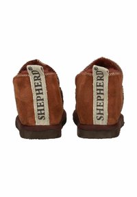 Un par de pantuflas de ante marrón con forro de lana y tableros de tela beige en el talón etiquetados "SHEPHERD", mostradas desde la parte trasera.