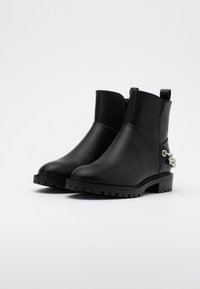 Botines de cuero negro con suela gruesa, que presentan un lazo lateral y un acento de cadena de metal. Textura lisa con detalles de costura mínimos.