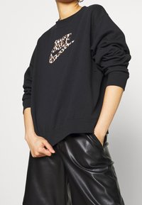 Zwarte sweatshirt met een leopardprint Nike-logo. Heeft een relaxte pasvorm, ronde halslijn en ribgebreide boorden. Gestyleerd met zwarte leren shorts.