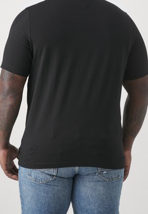 Mężczyzna w prostym czarnym t-shircie z krótkim rękawem i jasnoniebieskich dżinsach, pokazany od tyłu na jednolitym tle.