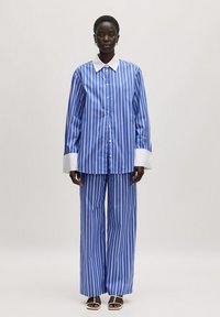 Blau-weiß gestreiftes, knöpfbares Hemd mit großem Kragen, kombiniert mit weiten Hosen. Glatte Stofftextur. Weiße Manschettenakzente.