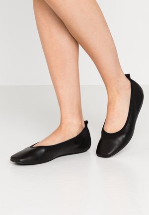 Jambes portant des ballerines en cuir noir avec des bouts carrés arrondis, posées sur une surface blanche lisse.