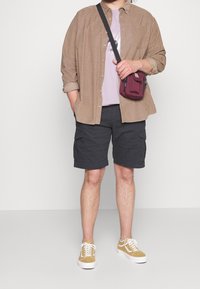 Camisa beige con botones sobre una camiseta lila, pantalones cortos de carga negros y zapatillas mostaza. Se lleva un pequeño bolso cruzado burdeos.