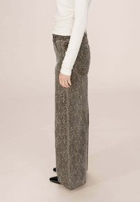 Personne portant un pantalon large à taille haute en imprimé léopard avec des poches, un haut blanc à manches longues et des chaussures noires à bout pointu, se tenant de profil.