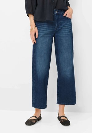 Kvinde iført blå brede, korte jeans, sorte Mary Jane-sko i mesh og sort bluse, står med den ene hånd i lommen.
