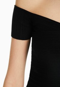 Gros plan sur le bras supérieur et l'épaule d'une personne portant un haut en tissu côtelé noir, à épaules dénudées, avec des manches courtes.