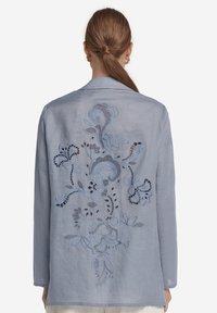 Ljust blå linneskjorta med intrikata blommösterbroderier och utskärningar på baksidan. Mjuk textur, avslappnad passform och långa ärmar.