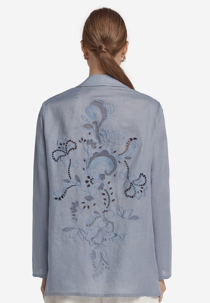 Ljust blå linneskjorta med intrikata blommösterbroderier och utskärningar på baksidan. Mjuk textur, avslappnad passform och långa ärmar.