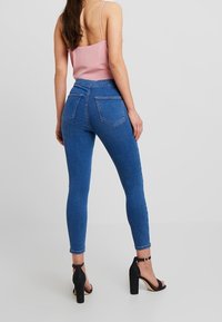 Woman wearing blue skinny jeans, pink sleeveless top, and black high-heeled sandals, standing against a plain light background.