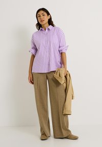 Camisa de botones de color púrpura claro con rayas blancas, mangas enrolladas y un bolsillo delantero, combinada con pantalones de pierna ancha beige y zapatos beige.