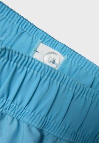 Costumi da bagno azzurri chiari con vita elasticizzata e chiusura con bottone. La texture è liscia, con sottili dettagli di cucitura orizzontale.