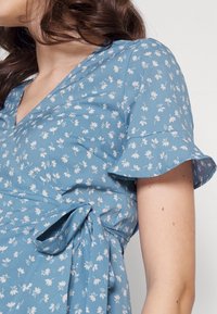 Blaues Blumenwickelkleid mit kurzen Rüschensleeves, aus leichtem Stoff gefertigt, der weiße Blumenmuster und ein seitliches Bindeband aufweist.