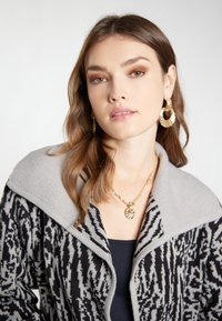 Strickjacke mit grauem Kragen und einem schwarzen und grauen gemusterten Oberteil; kombiniert mit goldenen Creolen und geschichteten goldenen Halsketten.