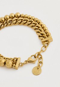 Topeltöödeldud kuldne käevõru, millel on poleeritud ümmargused ja ovalsest lingid, homaariklipp ja väike ümmargune graveeritud brändi ripats valgel taustal.