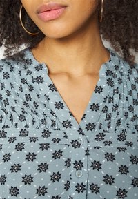 Blouse bleu clair avec un motif floral noir et des pois, fermeture à boutons, détails plissés au niveau de l'encolure et col court.