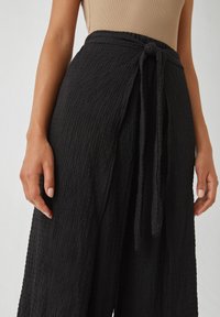 Pantalons larges noirs texturés avec une finition douce et froissée, dotés d'une taille nouée. Le tissu semble léger et respirant.