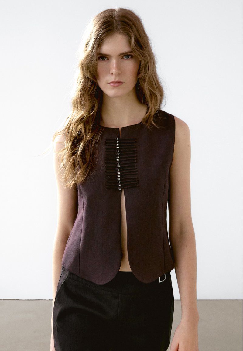 Mouwloze, donkerpaarse top met een voorste split en gestructureerde verticale accenten met metalen details. Glad materiaal, aansluitend ontwerp.