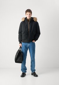 Geacă puf cu glugă căptușită cu blană, cu buzunare frontale; asortată cu blugi din denim albastri și pantofi din piele neagră; ținând o geantă neagră.
