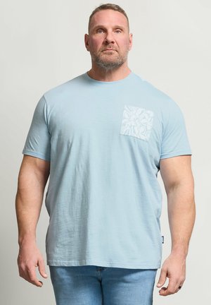 Muskularny mężczyzna stoi, ubrany w jasnoniebieską koszulkę z krótkim rękawem, z wzorzystą kieszenią na piersi i średnio niebieskie dżinsy na jednolitym tle.