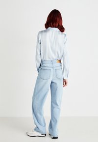 Camisa de cetim listrada azul-claro com mangas longas, combinada com jeans de denim claro de cintura alta e ténis brancos. Detalhe visível do bolso traseiro.