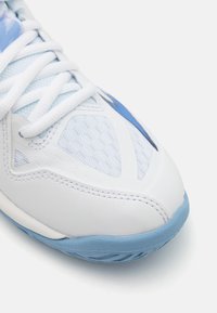 Chaussure de sport blanche avec une tige en mesh texturé, des accents et des lacets bleu clair, un bout arrondi et une semelle en caoutchouc bleu flexible.