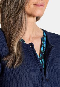 Frau mit langen braunen Haaren, die einen strukturierten marineblauen Cardigan über einer blau-grünen Bluse mit floralem Muster trägt, von den Schultern bis zum Kinn gezeigt.