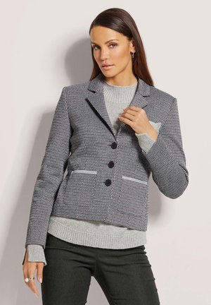 Hounds-tooth geblokte blazer in marineblauw en wit, met een notch-lapel, drie knopen en twee voorzakken. Gelaagd over een lichtgrijze trui.