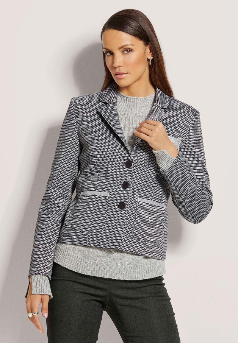 Blazer im Hahnentrittmuster in Marineblau und Weiß, mit Reverskragen, drei Knöpfen und zwei vorderen Taschen. Über einem hellgrauen Pullover getragen.