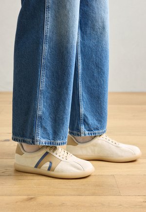 Beige suede sneakers with blue and brown accents, featuring a rounded toe, lace-up design, and a flexible tan rubber sole, worn with blue jeans.