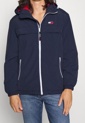 Homme portant une veste Tommy Jeans bleu marine avec une fermeture éclair blanche, une doublure de capuche rouge et un jean bleu, debout devant un fond uni.