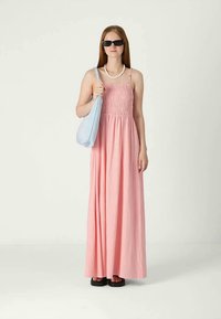 Mlada žena u ružičastoj maxi haljini, crnim sandalama, sunčanim naočalama, bijelom ogrlicom i s svijetloplavom torbom preko ramena, stoji.