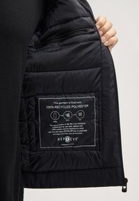Interno di un giubbotto puffa nero con design trapuntato e un'etichetta che dettaglia l'isolamento in poliestere riciclato al 100%. Include una tasca con zip.