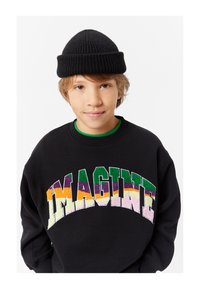 Felpa nera con lettering "IMAGINE" in rilievo e multicolore. Un berretto a coste nero è posato su capelli castano chiaro corti.