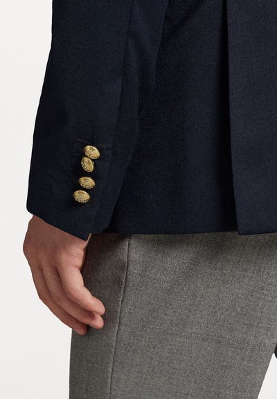 Manga de um blazer azul-marinho com quatro botões dourados, tecido texturizado e corte ajustado. Calças cinzas visíveis na parte inferior com um acabamento liso.