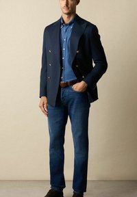 Man in een marineblauwe dubbelbreasted blazer met gouden knopen, blauw button-up shirt, blauwe jeans, een bruine riem en donkerbruine schoenen.