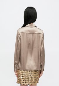 Jedwabista bluzka w kolorze taupe z długim rękawem, kołnierzykiem i plisą na plecach, zestawiona z błyszczącą złotą spódnicą z cekinów zdobioną warstwowymi kółkami.