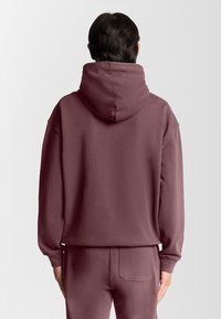 Mørkerød hættetrøje lavet af blødt stof med en stor hætte og dropped shoulders. Bagfra ses en afslappet pasform og minimal detaljer.
