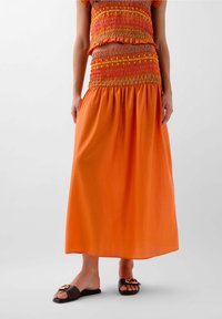 Oranges Smocked-Crop-Top mit bunten Nähten, kombiniert mit einem fließenden orangen Maxir Rock und braunen Rutschsandalen mit einer dekorativen Schnalle.
