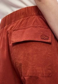 Gros plan sur une personne portant un pantalon de couleur rouille avec une taille élastique et une poche arrière ornée d'un patch logo crocodile.