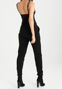 Black jumpsuit featuring a fitted bodice and loose pants, with logo-printed straps, elastic cuffs, and high-heeled ankle boots.