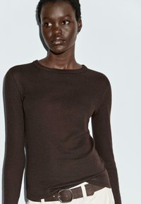Maglione lungo marrone a maniche lunghe realizzato in tessuto a maglia con scollo a girocollo. Il modello lo abbina a pantaloni chiari e una cintura abbinata.
