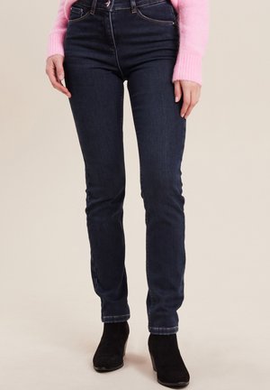 Jean skinny en denim bleu foncé avec taille haute, doté de cinq poches, de coutures discrètes et d'une texture lisse. Porté avec des bottines noires.
