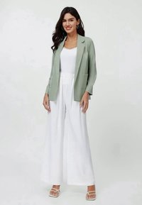 Blazer verde chiaro con revers, abbinato a un top bianco e pantaloni bianchi a gamba larga. Le scarpe trasparenti completano l'outfit.