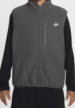 Uomo che indossa un gilet smanicato in pile grigio scuro con zip con logo Nike e una maglia nera a maniche lunghe, visibile dal collo ai fianchi.