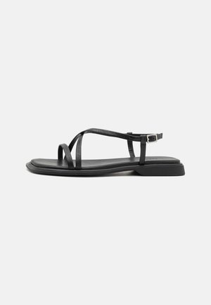Sort flad sandal med tynde remme, spænde og let hævet hæl på en ensfarvet lys baggrund.